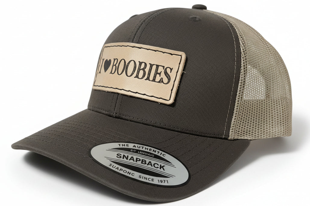 Black cap with 'I ❤️ Boobies' patch on a wooden surface