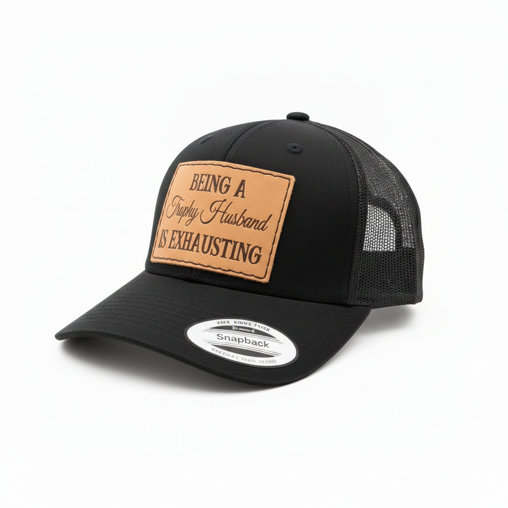 Black cap with a leather patch reading 'Being a Trophy Husband is Exhausting' on a cowhide rug background.