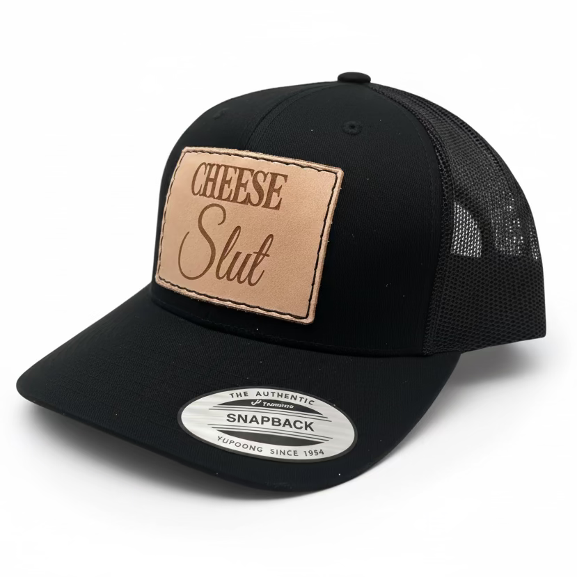 A black trucker hat with a tan leather patch on the front that reads 'CHEESE SLUT' in bold letters.