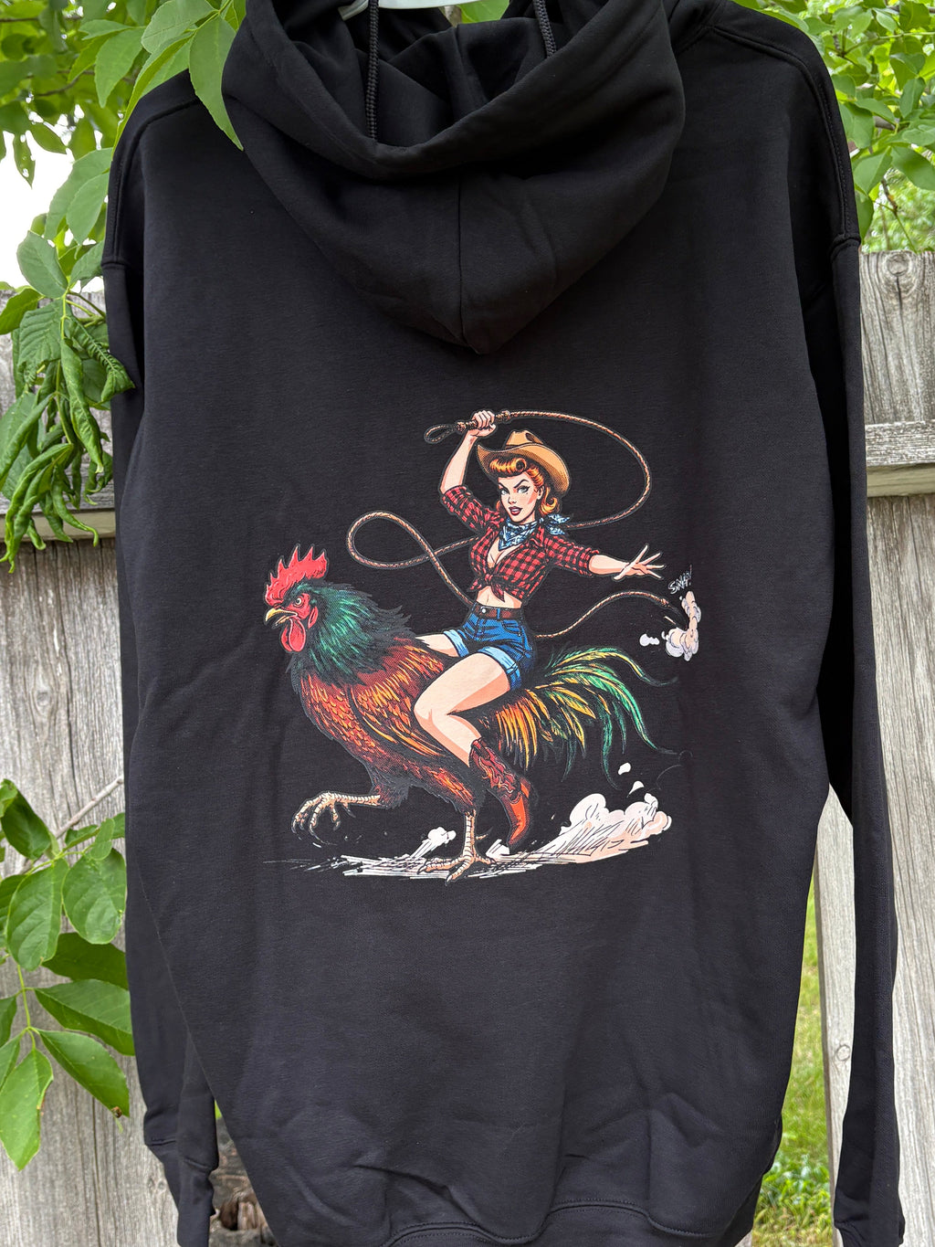 Black hoodie with a colorful graphic of a woman riding a rooster, hanging against a wooden fence with greenery.