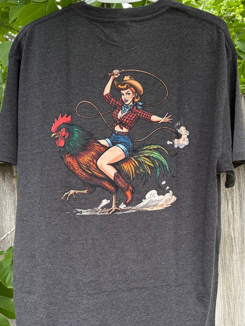 Gray t-shirt with a graphic of a woman on a rooster, hanging on a wooden fence with greenery in the background.