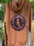 Brown hoodie with a logo on the back, hanging against a wooden fence and greenery background.