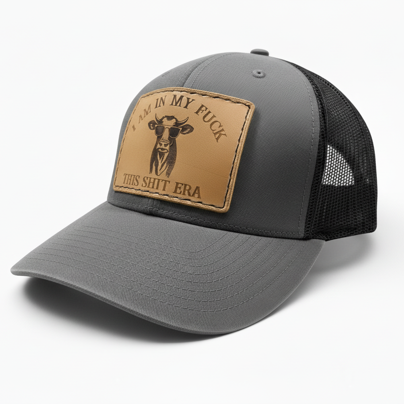 Gray and black cap with a brown leather patch featuring text and a cow illustration on a white background.