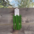 Green fringe earrings hanging from a white display card, attached to a weathered wooden fence with trees and sky in the background.