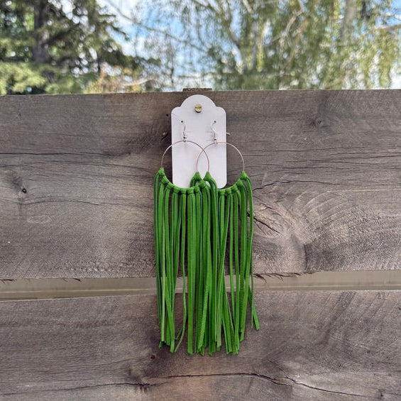 Green fringe earrings hanging from a white display card, attached to a weathered wooden fence with trees and sky in the background.