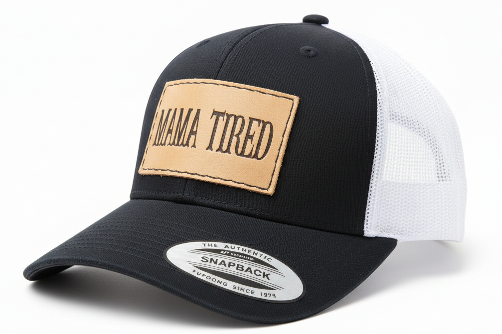 Black and white snapback cap with a tan patch labeled 'Mama Tired' on a white background