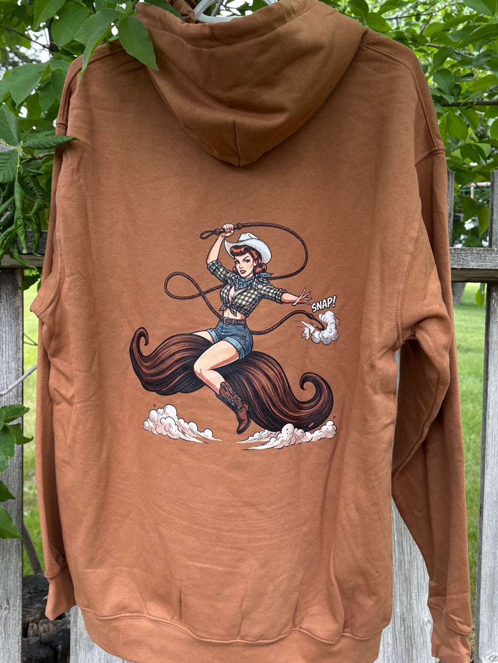 Brown hoodie with graphic design of a woman riding a moustache on a wooden fence with greenery in the background