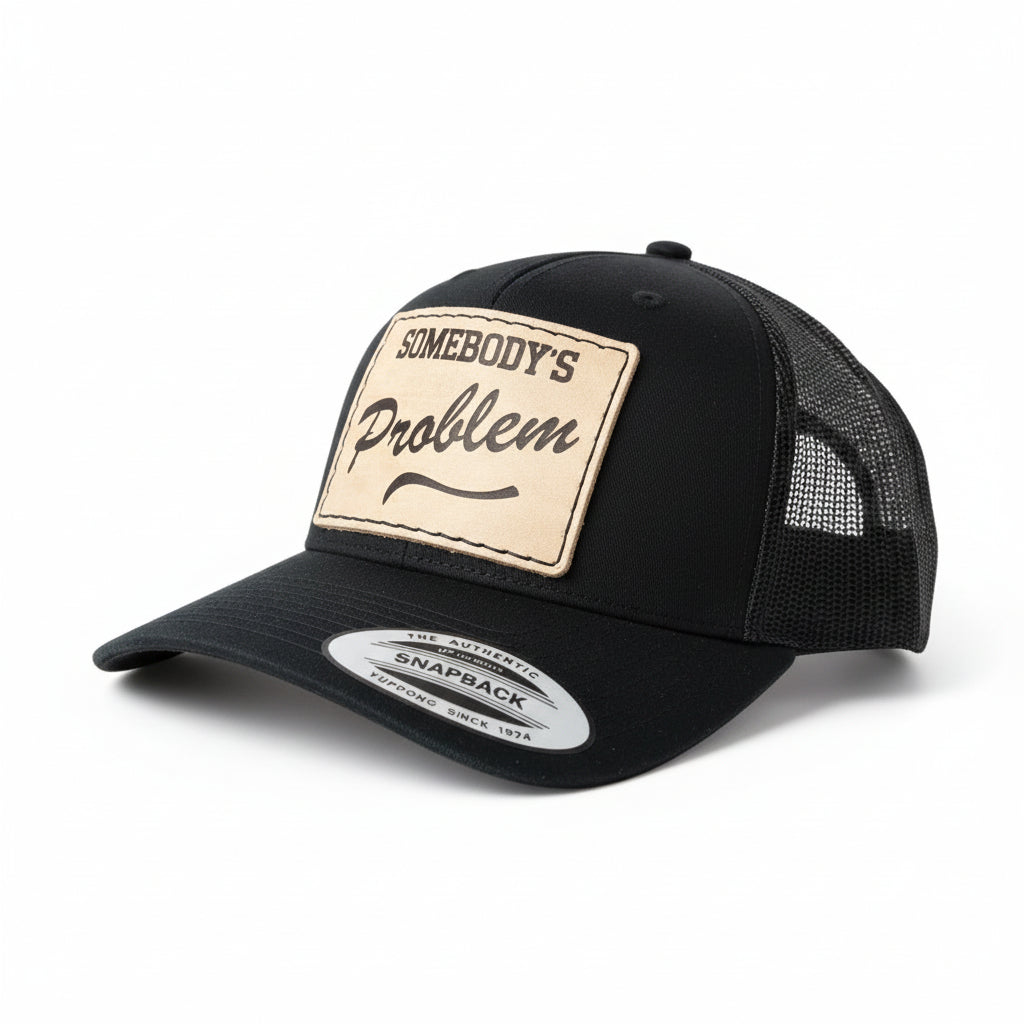 Black trucker cap with a beige patch displaying 'Somebody's Problem' on a white background