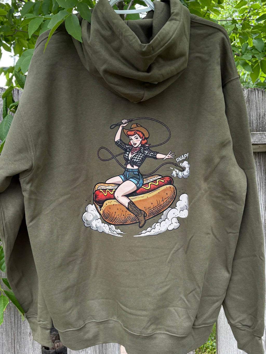Green hoodie with a graphic design of a person on a hot dog, hanging on a wooden fence with greenery in the background.