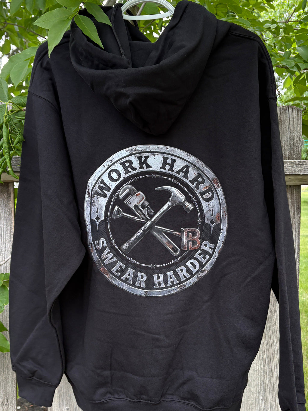 Black hoodie with 'Work Hard, Swear Harder' graphic on a wooden fence with greenery.