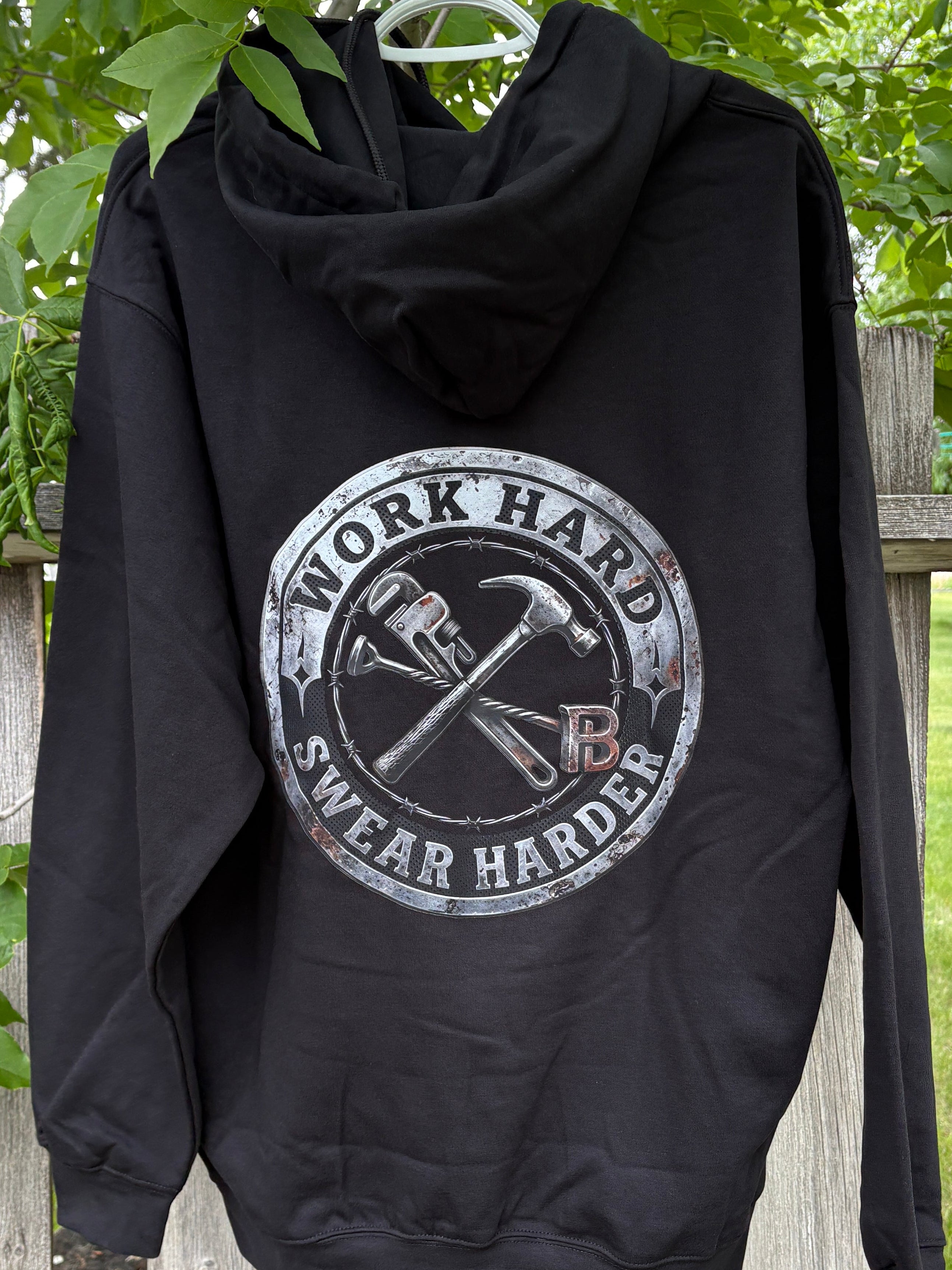 Black hoodie with 'Work Hard, Swear Harder' graphic on a wooden fence with greenery.