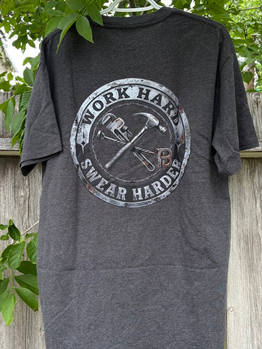 Gray t-shirt with 'Work Hard, Swear Harder' graphic on a hanger against a wooden fence and greenery background